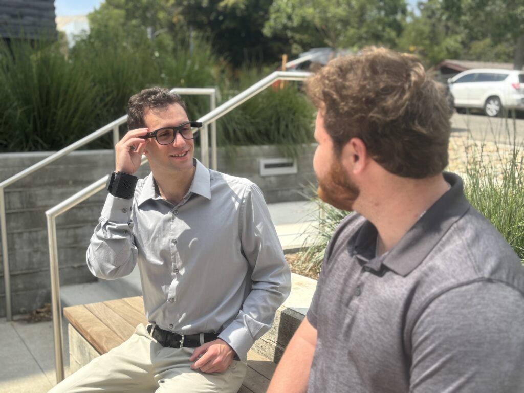 Man wearing ALEYE wristband and glasses conversing on a bench with another man.